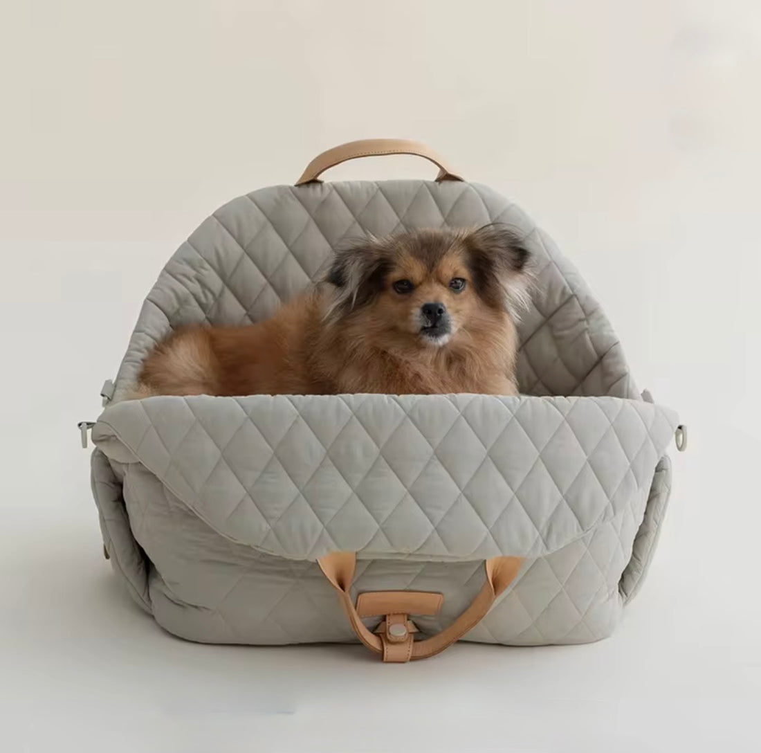 A small fluffy brown dog sits inside a light gray quilted pet car seat with tan leather handles, photographed against a plain off-white studio background.

